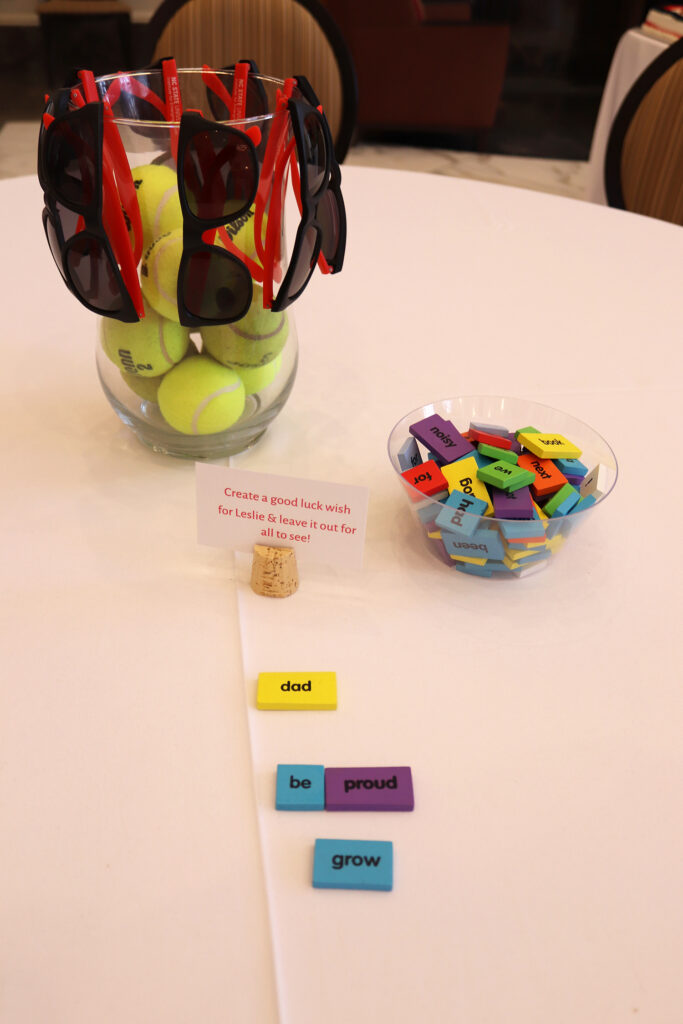 A table centerpiece of tennis balls and sunglasses in a vase, next to a glass bowl of colored tiles with various words printed on them.