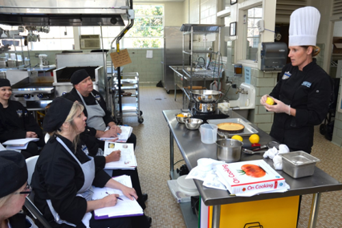 Four adult students in a culinary arts class watch the instructor at the head of the class.