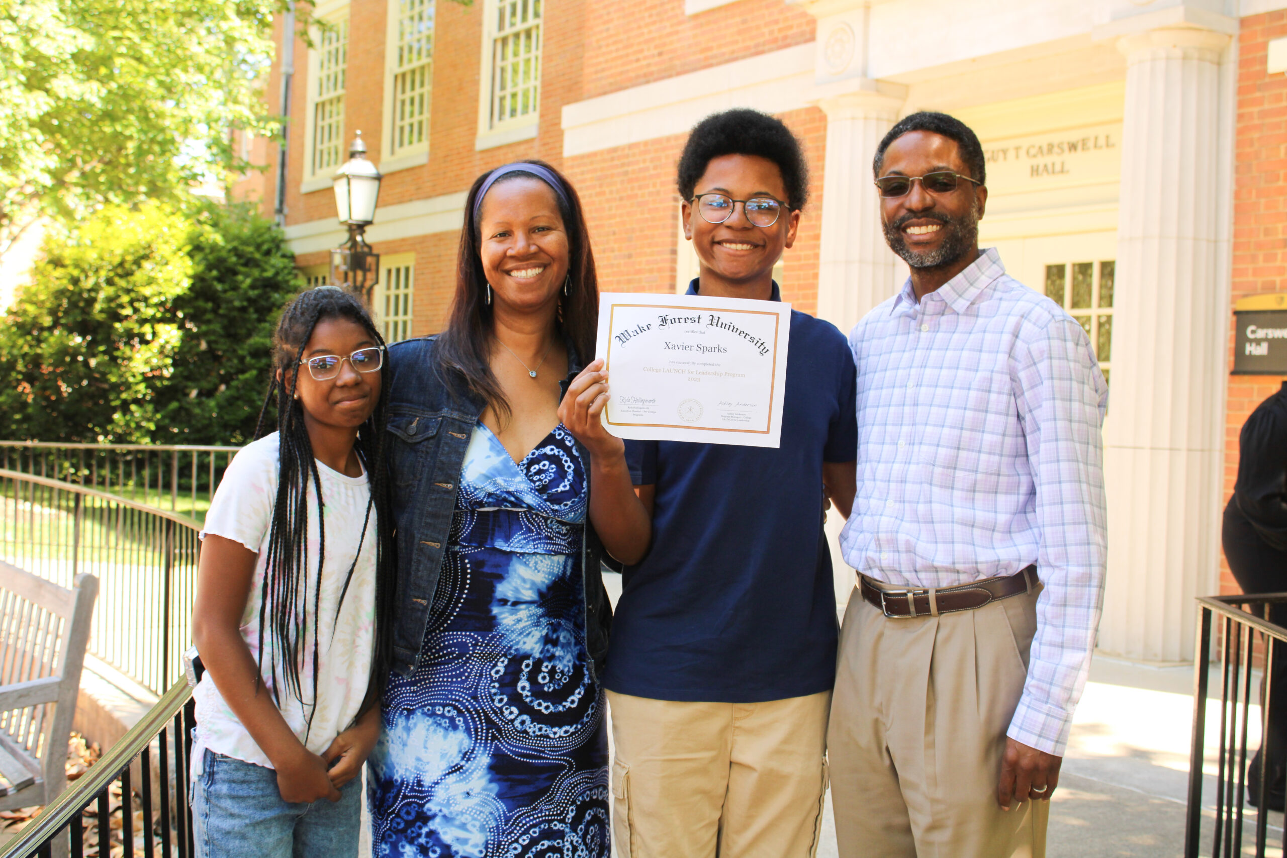 A family of four stands together. The teenage son is holding a certificate for completing a college prep program.