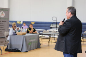 A man speaks into a microphone at the front of a room. In the background, several people sit at a table listening.