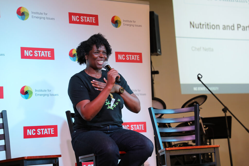 A woman sits on a tall stool at the front of an event room speaking into a microphone about nutrition