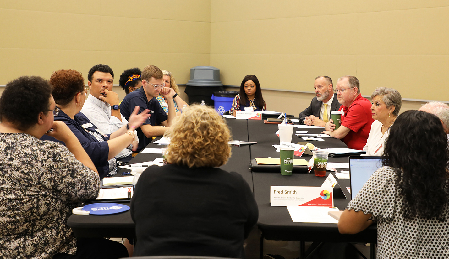 Photo of thirteen adults sitting around a large rectangular table having a meeting. One person on the left side of the table is talking and everyone else is looking at them, listening to what they are saying.