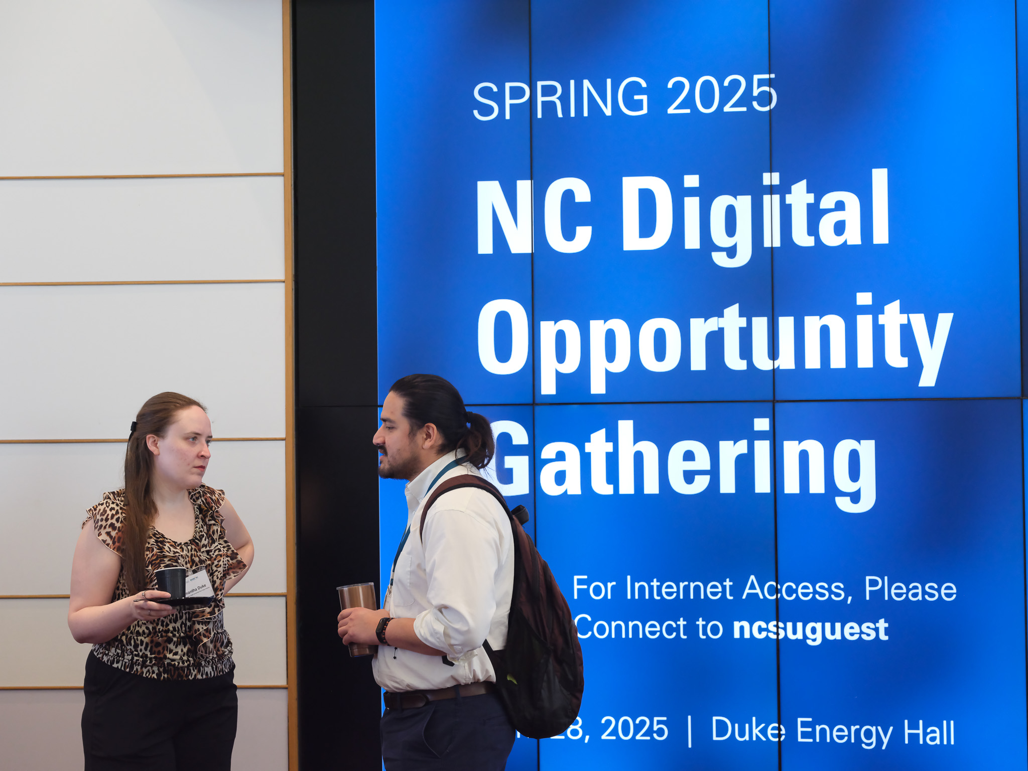A man and a woman talking in front of a large digital sign that says "NC Digital Opportunity Gatheirng'