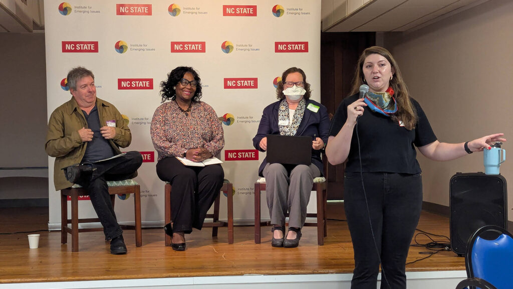 Two women and one man sitting in chairs on a stage. A third woman is standing in front of the stage, speaking into a microphone she is holding in her right hand. The three people on stage are looking at the woman who is speaking into the microphone. Behind the people on stage is a large banner with the logos for NC State University and the Institute for Emerging Issues repeated multiple times.