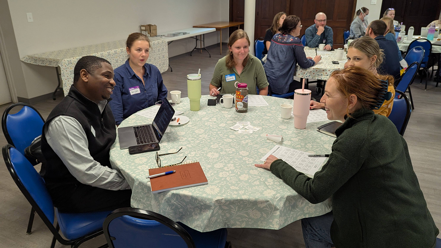 Five people sitting at a large table, talking with eachother. Two other large tables can be seen in the background with other people sitting around them and talking.